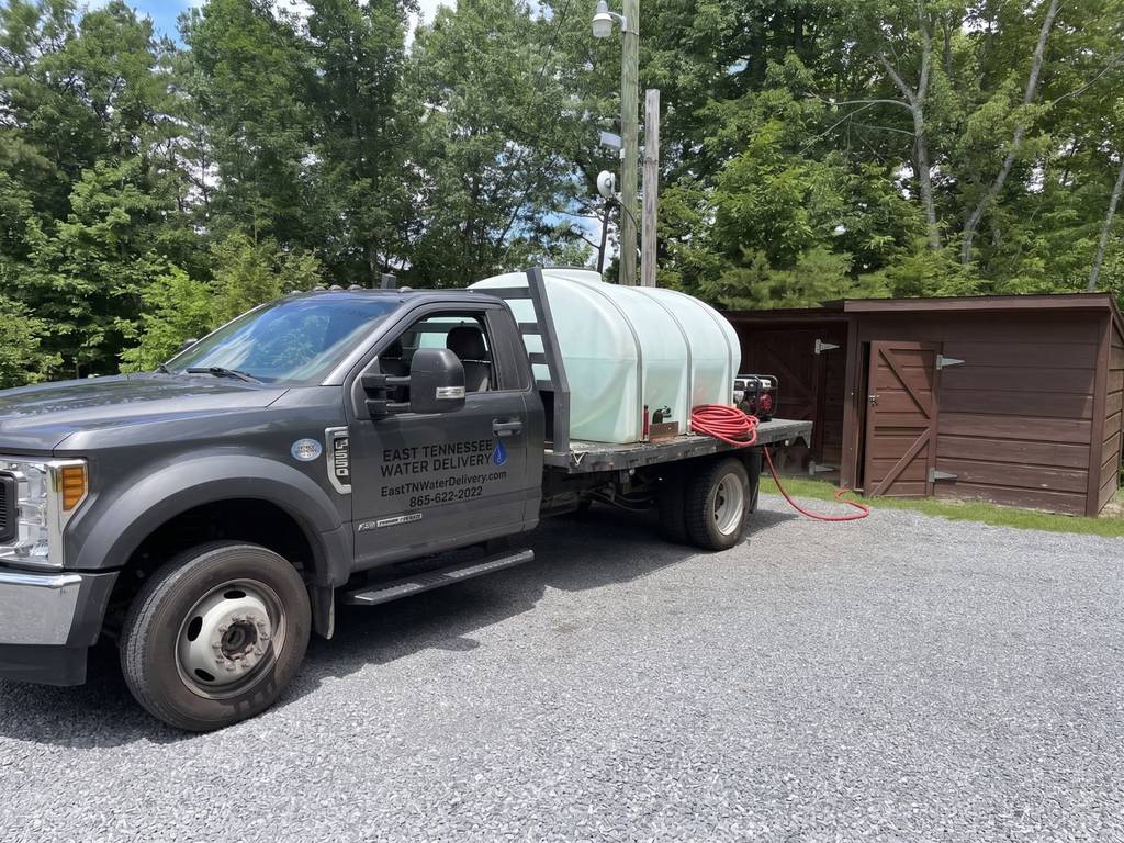 East Tennessee Water Delivery flatbed truck carrying a large white water tank, parked on a gravel driveway beside a wooden shed and green trees